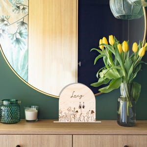 May include: A wooden sign with a mushroom and flower design. The sign is arched at the top and has the name "Lucy" engraved on it.