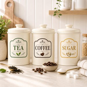 May include: Three cream-colored cylindrical canisters with lids, labeled TEA in green, COFFEE in brown, and SUGAR in gold. Each canister features a decorative border and matching illustrations of tea leaves, coffee beans, and sugar cubes.