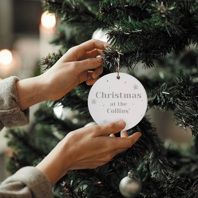 Peut inclure: Une d&eacute;coration de No&euml;l ronde et blanche avec le texte "Christmas at the Collins" en gris. L'ornement est orn&eacute; de petits flocons de neige et est plac&eacute; sur un sapin de No&euml;l vert. L'ornement est tenu par une personne.