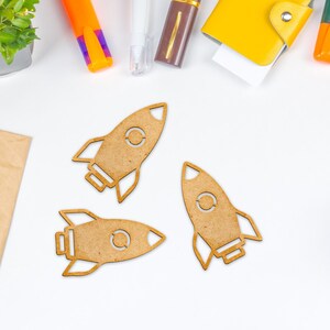 May include: Three brown wooden rocket ship cutouts on a white background. The rockets are arranged in a triangular pattern.