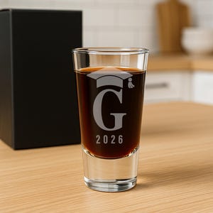 K&ouml;nnte beinhalten: Ein klares Schnapsglas, gef&uuml;llt mit einer dunklen Fl&uuml;ssigkeit, mit einem eingravierten Abschluss-Hut, dem Buchstaben "G" und dem Jahr "2026". Im Hintergrund befindet sich eine schwarze Box, und das Glas steht auf einer Holzoberfl&auml;che.