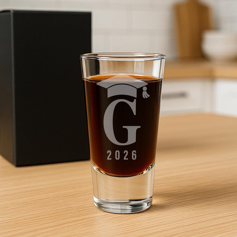 K&ouml;nnte beinhalten: Ein klares Schnapsglas, gef&uuml;llt mit einer dunklen Fl&uuml;ssigkeit, mit einem eingravierten Abschluss-Hut, dem Buchstaben "G" und dem Jahr "2026". Im Hintergrund befindet sich eine schwarze Box, und das Glas steht auf einer Holzoberfl&auml;che.