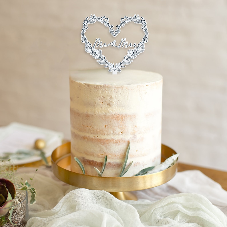 May include: A white wedding cake with a simple buttercream frosting and a delicate floral heart topper that says "Mr & Mrs". The cake is on a gold cake stand.