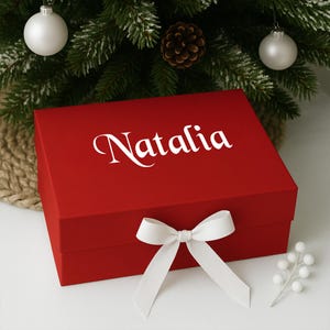 May include: A red gift box with the name "Natalia" in white script on the lid. A white bow is tied around the box. The box is in front of a Christmas tree with ornaments and a pinecone.