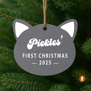 May include: A gray cat-shaped Christmas ornament with white accents and the text "Pickles' FIRST CHRISTMAS - 2025 -" in white. The ornament hangs from a natural twine string against a backdrop of green Christmas tree branches.