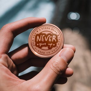 May include: A copper-coloured round token with the words "One step closer to your goal" and "Never give up" in a cloud shape. The token is held in a hand.