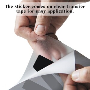Pode incluir: Uma m&atilde;o descolando uma fita de transfer&ecirc;ncia transparente de um adesivo. O adesivo tem um desenho preto.