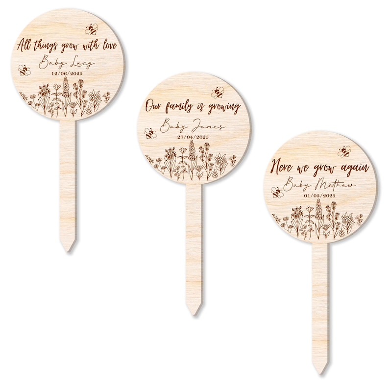 K&ouml;nnte beinhalten: Drei h&ouml;lzerne Gartenpf&auml;hle mit runden Schildern, die mit Blumenmustern und Bienenillustrationen verziert sind. Die Schilder tragen personalisierte Botschaften f&uuml;r Neugeborene, darunter "All things grow with love Baby Lacy 12/06/2025", "Our family is growing Baby James 27/04/2025" und "Never we grow again Baby Mathew 01/05/2025".