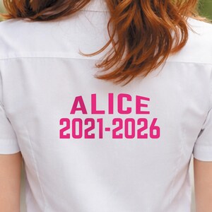 Puede incluir: Camisa blanca abotonada con el nombre "ALICE" y los a&ntilde;os "2021-2026" impresos en rosa brillante en la espalda. La camisa es de tela lisa. La imagen muestra la parte trasera de la camisa.