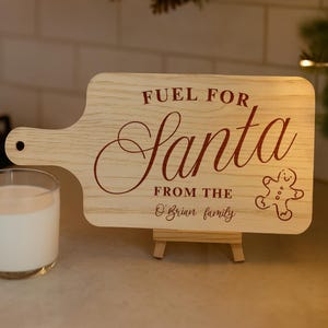 May include: A wooden cutting board with a handle, featuring the text "FUEL FOR Santa" in red script, along with a gingerbread man illustration. The text "FROM THE O'Brian family" is also included. A glass of milk is visible on the left.
