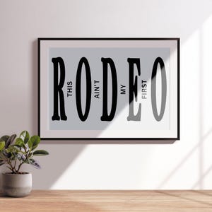 May include: Black and white typography print with the word "RODEO" in large letters. The text "This ain't my first" is written vertically within the letters.