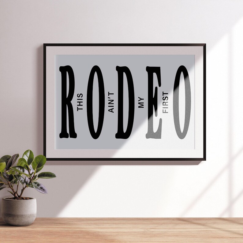 May include: Black and white typography print with the word "RODEO" in large letters. The text "This ain't my first" is written vertically within the letters.