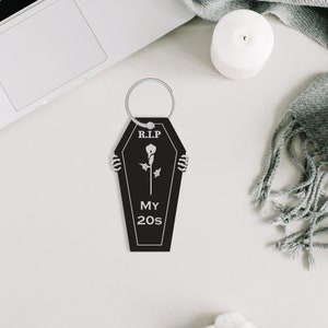 May include: Black coffin-shaped keyring with a silver ring. The keyring has a white rose and the text "R.I.P. My 20s" printed on it.