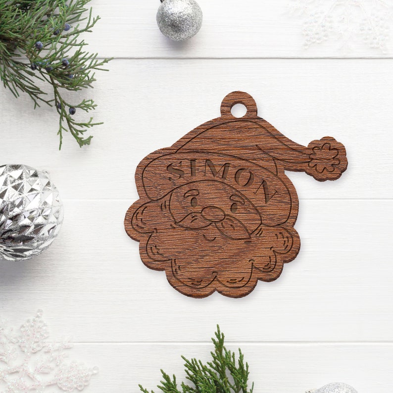 May include: A wooden Christmas ornament shaped like Santa Claus's face with the name "Simon" carved into the hat. The ornament is brown and has a rustic look.