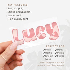 May include: A pink and white sticker with the name "Lucy" in a bubble font. The sticker is held by a hand. The text on the sticker says "Easy to apply", "Strong and durable", "Waterproof", and "High-quality print".