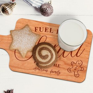 May include: A wooden serving board with the text "FUEL the soul" and "Andrews family" engraved on it. The board holds a star-shaped cookie, a swirl cookie, a glass of milk, and a gingerbread man cookie. The board is light brown.
