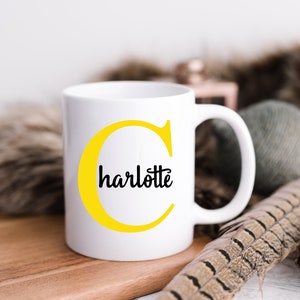 K&ouml;nnte beinhalten: Wei&szlig;e Keramiktasse mit einem gelben Buchstaben C und dem Wort "charlotte" in schwarzer Schrift.