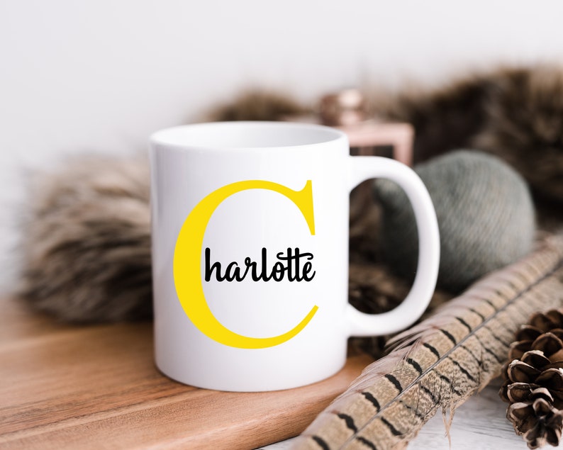 K&ouml;nnte beinhalten: Wei&szlig;e Keramiktasse mit einem gelben Buchstaben C und dem Wort "charlotte" in schwarzer Schrift.