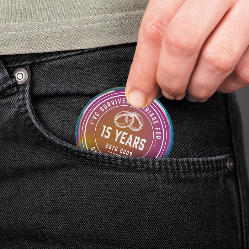 Puede incluir: Un recuerdo redondo e iridiscente con el texto "I've survived marriage for... 15 years ESTD 2009" y dos anillos entrelazados. El art&iacute;culo se sostiene en un bolsillo de unos vaqueros negros.