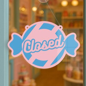 May include: A pink and blue candy-shaped sign with the word "Closed" in cursive script. The sign is hanging on a glass door, indicating the business is not open. The sign has a candy wrapper design.