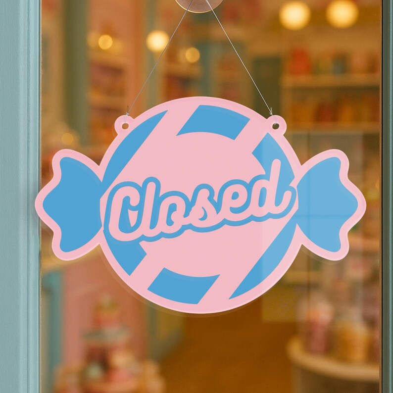 May include: A pink and blue candy-shaped sign with the word "Closed" in cursive script. The sign is hanging on a glass door, indicating the business is not open. The sign has a candy wrapper design.