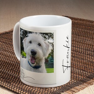 May include: White ceramic mug featuring a photo of a smiling white dog with a blue collar. The mug has the name "Frankie" in cursive script on the side and a paw print graphic. The mug is on a brown woven mat.