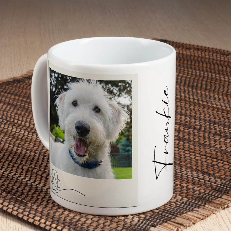 May include: White ceramic mug featuring a photo of a smiling white dog with a blue collar. The mug has the name "Frankie" in cursive script on the side and a paw print graphic. The mug is on a brown woven mat.