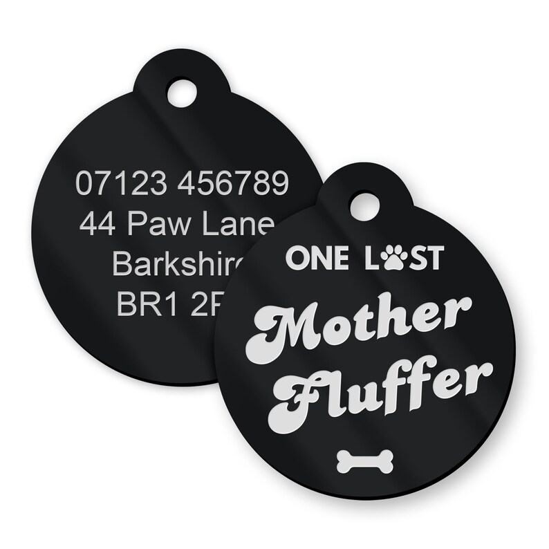 Op de afbeelding: Twee zwarte ronde huisdier-ID-tags. De grotere tag heeft de tekst "ONE LOST" en "Mother Fluffer" in wit met een pootprint en een beensymbool. De kleinere tag heeft de tekst "07123 456789", "44 Paw Lane", "Barkshire" en "BR1 2F" in wit.