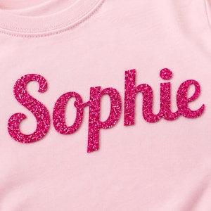 Puede incluir: Una camiseta rosa claro con el nombre "Sophie" en letras grandes y brillantes de color rosa intenso. Las letras est&aacute;n en cursiva y centradas en la camiseta. La camiseta est&aacute; hecha de un material suave.