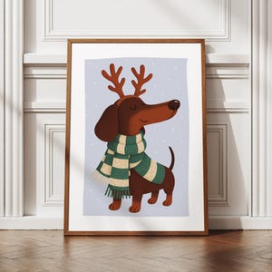 May include: A framed print featuring a cartoon dachshund wearing a green and white scarf and reindeer antlers. The dog is brown with a long snout and is set against a light blue background with white dots.