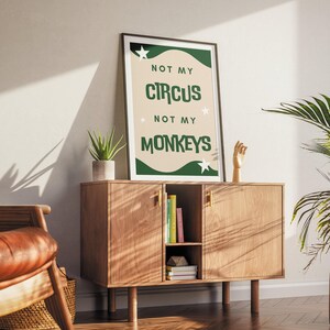 May include: A green and white poster with the text "Not my circus, not my monkeys" on a beige background. The poster is framed and hanging on a wall above a wooden cabinet.