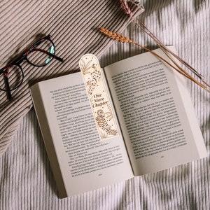 Op de afbeelding: Houten bladwijzer met een lasergesneden ontwerp met een luipaard en een tijger, met de tekst "One More Chapter". De bladwijzer ligt in een open boek, met een bril en een beige deken.