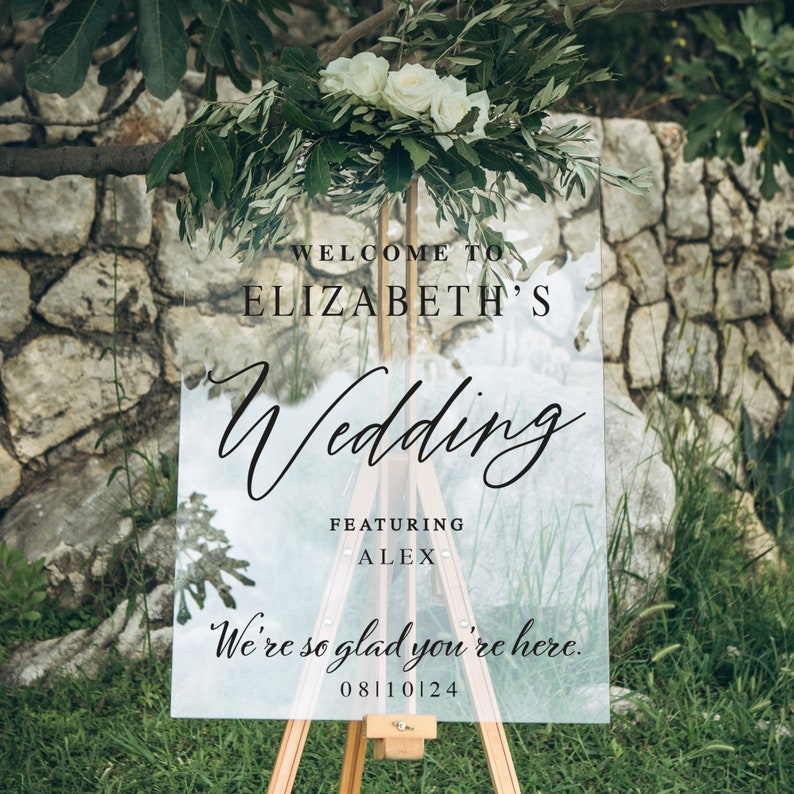 K&ouml;nnte beinhalten: Ein klares Acryl-Hochzeitschild mit dem Text "Willkommen zur Hochzeit von Elizabeth und Alex. Wir freuen uns, dass Sie da sind. 08110124". Das Schild ist mit wei&szlig;en Blumen und Gr&uuml;npflanzen verziert.