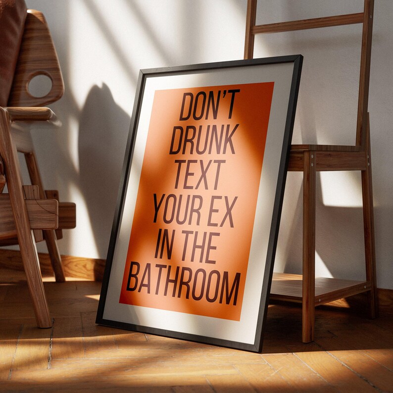 Op de afbeelding: Een ingelijst print met een oranje achtergrond en zwarte tekst die "Drink niet en sms je ex niet in de badkamer" zegt.