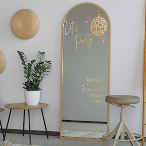 May include: A tall, arched mirror with a wooden frame. The mirror has a gold-colored vinyl decal with the text "Let's Party" and a disco ball hanging from it. The text "Welcome to Frankie's Birthday Party!" is also on the mirror.