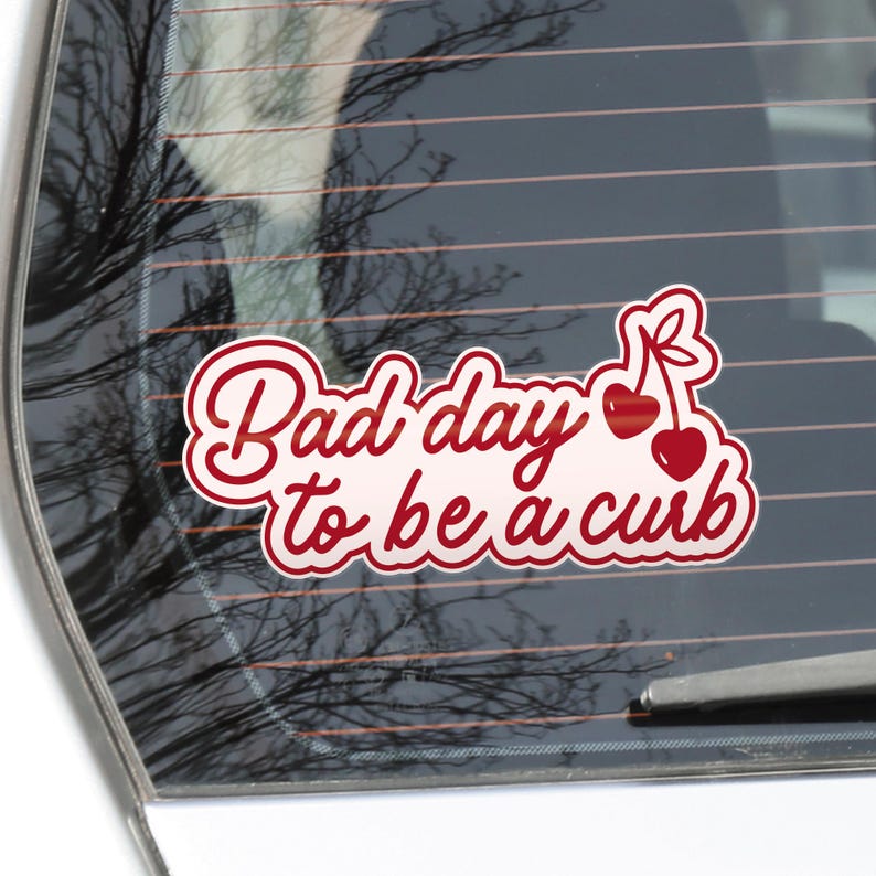 K&ouml;nnte beinhalten: Wei&szlig;er Aufkleber mit rotem Schreibschrift-Text: &bdquo;Bad day to be a curb&ldquo;. Zwei rote Kirschen mit gr&uuml;nen Stielen rechts. Der Aufkleber befindet sich auf einer Autoscheibe.