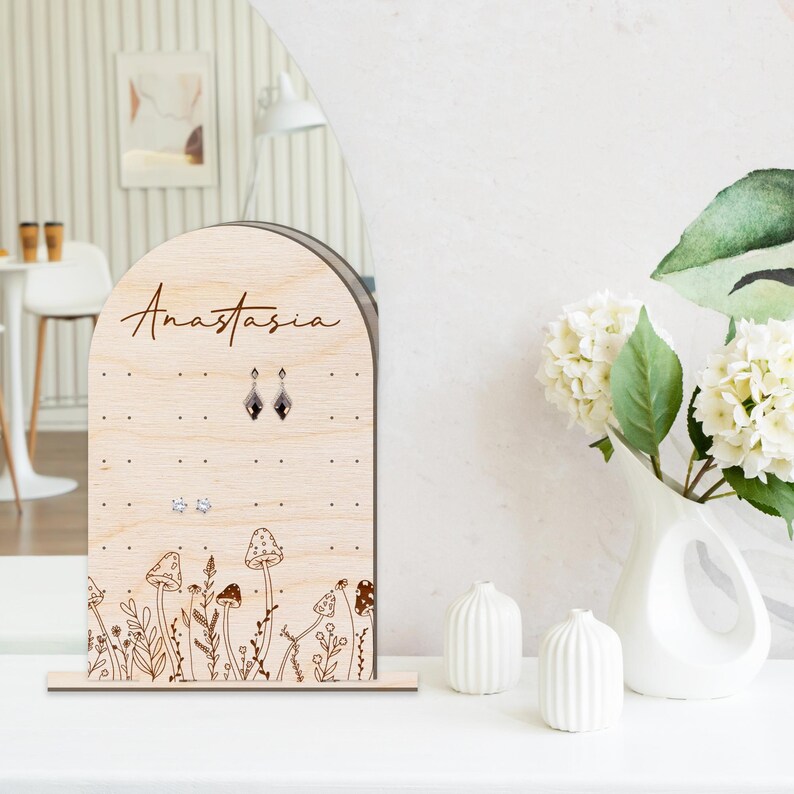 May include: Wooden earring display stand with the name Anastasia engraved on the front. The stand features a mushroom and floral design and has multiple holes for earrings.