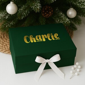 May include: A dark green gift box with the name "Charlie" in gold lettering. The box is tied with a white ribbon bow. The background includes a Christmas tree with ornaments and a pine cone.