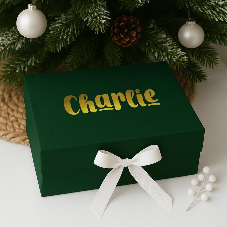 May include: A dark green gift box with the name "Charlie" in gold lettering. The box is tied with a white ribbon bow. The background includes a Christmas tree with ornaments and a pine cone.