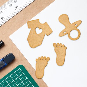 May include: Five wooden cutouts in the shape of a baby onesie, a pacifier, and two footprints. The onesie has a heart cutout in the center. The cutouts are light brown and have a rough texture.