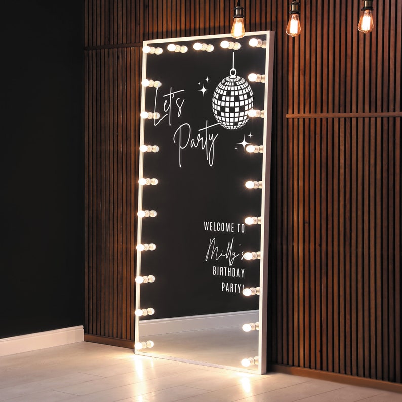May include: A white, full-length mirror with a white frame and a black background. The mirror has a row of light bulbs around the perimeter. The mirror has a white vinyl decal with the text "Let's Party" and a disco ball graphic. The mirror also has the text "Welcome to Milly's Birthday Party".