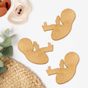 May include: Three wooden cutouts of a fetus in a fetal position, with a light brown color. The cutouts are on a white surface, along with a pacifier and a woven basket.