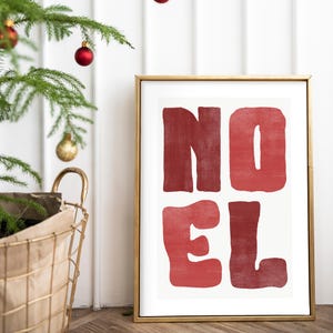 May include: A framed print with the word "NOEL" in large, red, block letters. The artwork is in a gold frame and is set against a white background. A Christmas tree and basket are in the background.