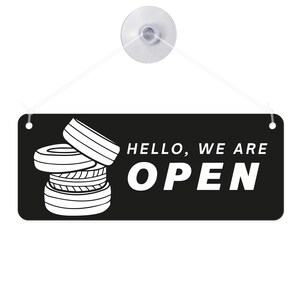 May include: Black rectangular sign with the words "HELLO, WE ARE OPEN" in white. A stack of tires is on the left side of the sign. The sign is suspended by a clear suction cup and string.