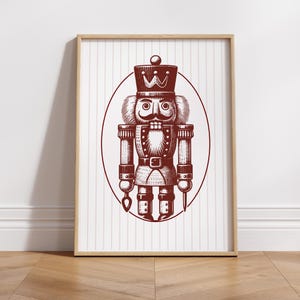 May include: A framed print featuring a detailed illustration of a nutcracker soldier in a maroon color. The nutcracker is depicted in a classic uniform with a tall hat, mustache, and holding tools. The background has vertical stripes.