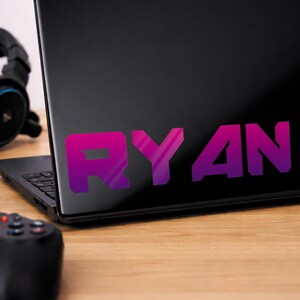 May include: A black laptop displaying the name "RYAN" in a purple and pink gradient. A black gaming controller and headphones are on a wooden surface.