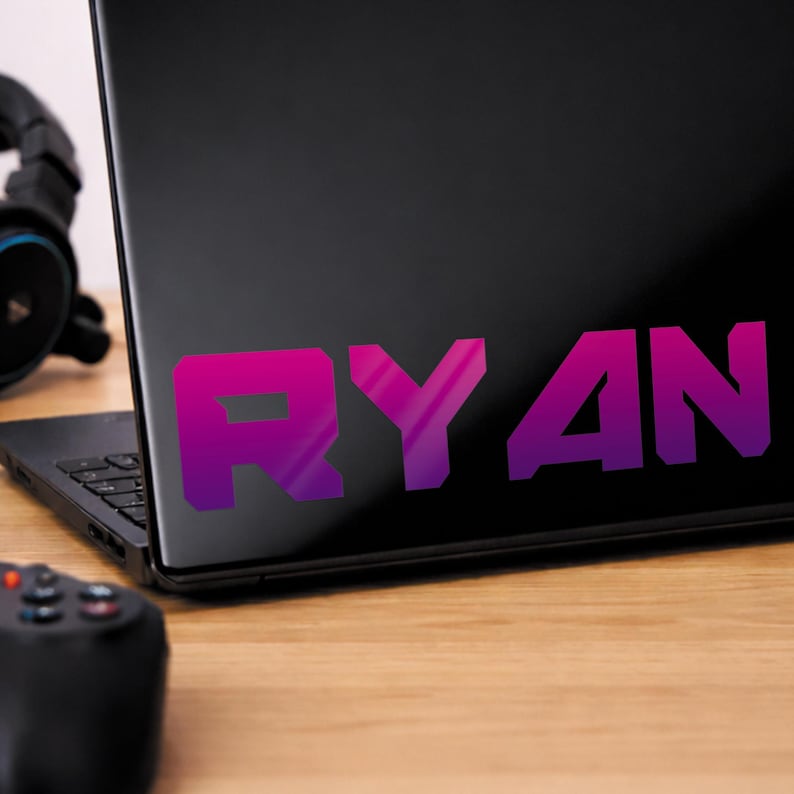 May include: A black laptop displaying the name "RYAN" in a purple and pink gradient. A black gaming controller and headphones are on a wooden surface.