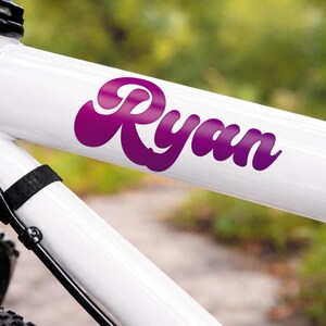 May include: A close-up of a white bicycle frame with the name "Ryan" in a purple, cursive font. The name is a sticker or decal, and the bicycle frame is angled, with a blurred green background.