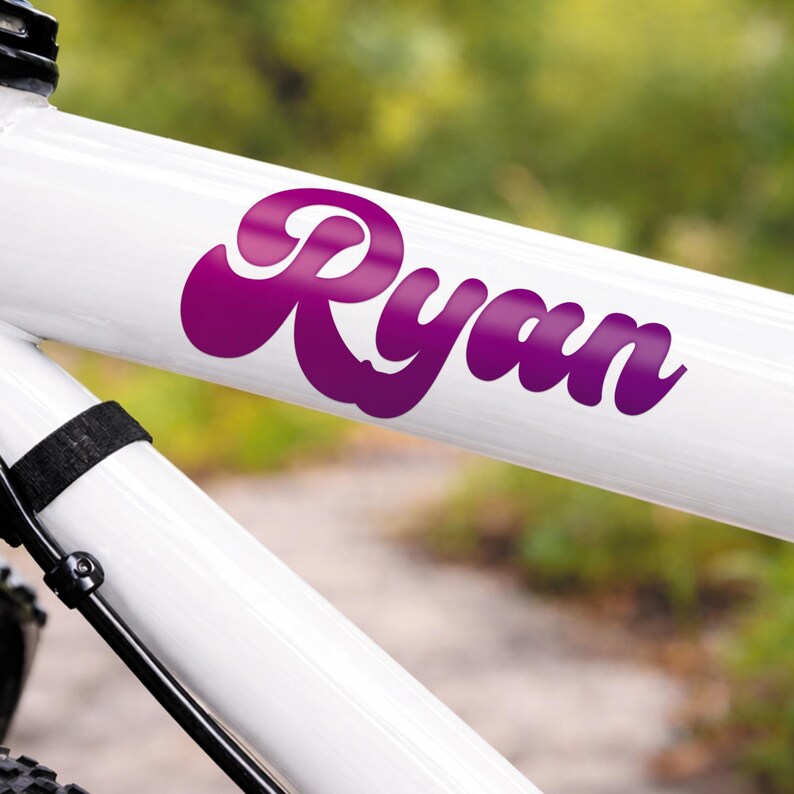 May include: A close-up of a white bicycle frame with the name "Ryan" in a purple, cursive font. The name is a sticker or decal, and the bicycle frame is angled, with a blurred green background.