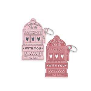 May include: Two pink and red slot machine keychains with the text "I hit the JACKPOT WITH YOU" in a white outline. The keychains are decorated with hearts and a small white ribbon.